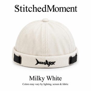Könnte beinhalten: Eine milchig weiße, schirmlose Mütze mit dem Text StitchedMoment oben und Milky White unten. Die Mütze hat eine schwarze, gestickte Haigrafik auf der Vorderseite, schwarze Seitenriemen mit Metallringen und einen zentralen Knopf auf der Krone.