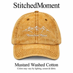 May include: A mustard yellow washed cotton baseball cap featuring an embroidered white mountain range graphic and the text Mountain Mama. The cap has a curved brim, stitched eyelets, and a button top, presenting a distressed texture.
