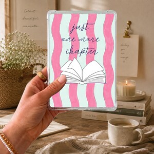 May include: A clear tablet case featuring a vertical wavy pink and light blue striped pattern. The design includes a white line-art illustration of an open book and the dark blue text "just one more chapter" centered on the front surface.