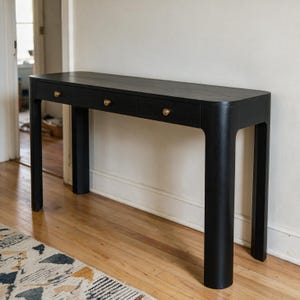 May include: A black wooden console table with a smooth rectangular top and rounded corners. The table features three horizontal drawers, each equipped with a single small circular brass knob. It stands on four sturdy, thick, curved legs.