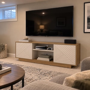 May include: A long wooden media console featuring two white cabinet doors with a geometric triangle pattern, an open center shelf holding a gaming console and controller, and a flat top surface supporting a large wall-mounted television.