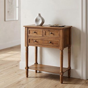 May include: A wooden console table featuring two small upper drawers and one wider lower drawer, all equipped with dark round knobs. The table stands on four turned legs with a lower shelf and holds a white ceramic vase and a small metal tray.