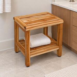 May include: A rectangular wooden shower stool with a slatted top and a lower shelf. The stool features a natural light brown wood grain finish and four sturdy legs. A folded white towel rests on the bottom shelf of the wooden furniture piece.