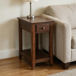 May include: A rectangular dark brown wooden end table featuring a single drawer with a small round metal knob and an open lower shelf. The table has four straight legs and a flat top surface that supports a silver lamp with a white shade.