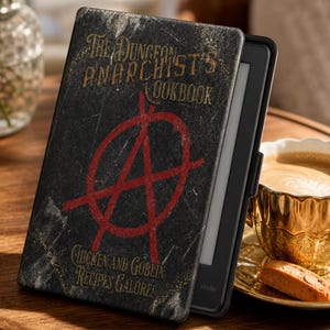 May include: A black e-reader case featuring the text The Dungeon Anarchist's Cookbook and Chicken and Goblin Recipes Galore in gold lettering, with a large red anarchy symbol centered on the cover, displaying a distressed, scratched texture.