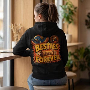May include: A black hooded sweatshirt featuring a graphic on the back with a blue dinosaur, a brown creature, and a severed arm. The text reads BESTIES FOREVER in yellow block letters against a red and orange sunburst background design.