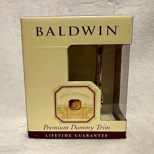 May include: A Baldwin brand premium dummy trim product in a rectangular cardboard box. The packaging is cream-colored with dark red accents, featuring the text Baldwin, Timeless Craftsmanship, and Premium Dummy Trim with a Lifetime Guarantee.