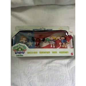 May include: A Cabbage Patch Kids Olympikids collectible set in original packaging featuring four small figures representing track and field, weightlifting, tennis, and gymnastics. The box displays a US flag background and the text Olympikids.