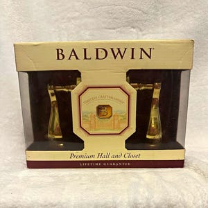 May include: A Baldwin brand Premium Hall and Closet door hardware set in a gold finish, packaged in a rectangular box with a clear plastic window. The box features the text Baldwin, Timeless Craftsmanship, and Lifetime Guarantee.
