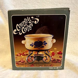 May include: A Country Corner brand box featuring a beige ceramic pot with two handles and a blue heart-shaped floral pattern. The pot sits on a black metal stand with a tea light candle inside. The box displays the text Country Corner and ITEM NO. 3553.