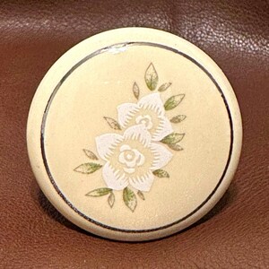 May include: A round, cream-colored ceramic cabinet knob featuring a central floral design. The graphic depicts two white flowers with detailed petals surrounded by small, muted green leaves. A thin, dark circular line borders the edge of the knob.