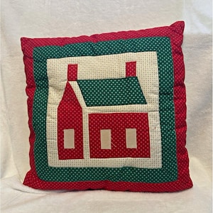 May include: A square quilted throw pillow featuring a central applique of a house with a red roof and walls, set against a cream background with a small geometric pattern. The pillow is framed by a green border and a red outer edge.