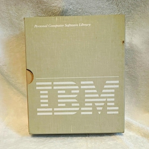 May include: A rectangular, light olive-green IBM Personal Computer Software Library box. The front features the white IBM logo composed of horizontal stripes, with the text Personal Computer Software Library printed in white above the logo.