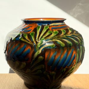 Peut inclure: Un vase en céramique rond présentant un vernis brillant avec un motif vibrant de feuilles orange, bleues et vertes sur un fond brun foncé. Le vase possède un corps large et bulbeux qui se rétrécit vers une ouverture circulaire étroite au sommet.