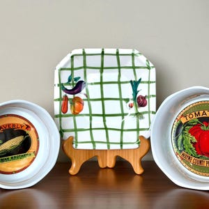 Puede incluir: Dos cuencos de cerámica blanca y un plato cuadrado. El cuenco izquierdo presenta un gráfico de maíz con el texto WAVERLY SEEMAN BROTHERS. El cuenco derecho muestra un gráfico de tomate con el texto TOMATOES WAYNE COUNTY PRESERVING CO. El plato tiene una cuadrícula verde.