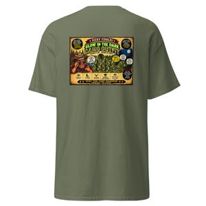 May include: An olive green t-shirt featuring a rectangular graphic on the back. The print displays a cartoon character in a hat, camouflage clothing, and text including "Dicky Fiddles Glow in the Dark Camo Outfit" and "Hunt. Fish. Camp. Disappear."