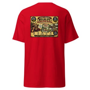 May include: A bright red short-sleeve t-shirt featuring a rectangular graphic on the back. The graphic displays a cartoon character in a hat, a book titled "Dicky Fiddles Wildlife Translator Manual," and various forest animals with speech bubbles.