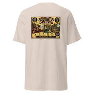 May include: A beige short-sleeve t-shirt featuring a large rectangular graphic on the back. The design depicts a cartoon hunter and wildlife with the text "Dicky Fiddles Invisible Decoys" and "Sometimes the best decoy is no decoy" in a vintage style.