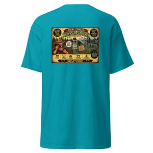 May include: A bright teal short-sleeve t-shirt featuring a rectangular graphic on the back. The graphic depicts a cartoon character in a hat, a spray bottle labeled "Dicky Fiddles Invisa Spray," a deer, and various text including "Spray Responsibly."