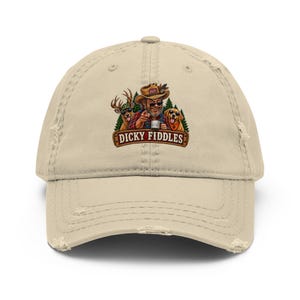 May include: A beige distressed baseball cap featuring a graphic of a person in a hat and sunglasses pointing forward, accompanied by a deer and a dog, with the text "DICKY FIDDLES" printed on a wooden-style banner below the illustration.