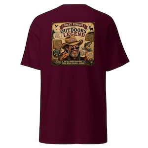 May include: A maroon short-sleeve t-shirt featuring a graphic on the back. The design shows a bearded person in a hat and sunglasses holding a mug, with text including "DICKY FIDDLES THE OUTDOORS LEGEND" and "NOT ALL HEROES WEAR CAPES... SOME WEAR CAMO & CLOGS!"