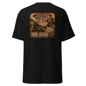 May include: A black t-shirt featuring a graphic print on the back. The design resembles a missing person poster with the text 'Dicky Fiddles Bigfoot Stole My ATV!' and includes an illustration of a person in a hat and a creature riding an ATV.