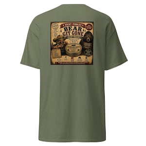 May include: An olive green t-shirt featuring a vintage-style graphic on the back. The design depicts a person in a cowboy hat pointing forward, a bear, and a tin labeled Dicky Fiddles Bear Git Gone Ointment, surrounded by various icons and text.