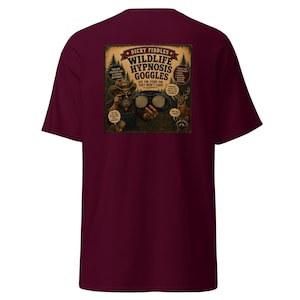 May include: A maroon short-sleeve t-shirt featuring a graphic on the back. The print depicts a person in a hat holding goggles with spiral lenses, surrounded by forest animals and text reading "Dicky Fiddles Wildlife Hypnosis Goggles".