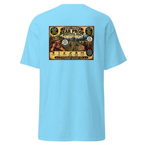 May include: A light blue t-shirt featuring a rectangular graphic on the back. The graphic depicts a person in a hat, a person in a camouflage suit, and a bear, with the text "Dicky Fiddles Bear Proof Survival Suit" and various labels.