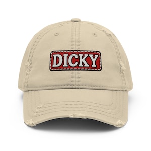May include: A beige distressed baseball cap featuring a rectangular patch on the front. The patch has a red background with a white border and the word DICKY embroidered in white capital letters across the center of the patch.
