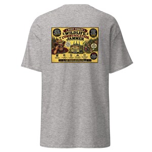 May include: A heather gray short-sleeve t-shirt featuring a rectangular graphic on the back. The design displays a vintage-style advertisement for a Dicky Fiddles Wildlife Communication Jammer, including illustrations of a person in a hat and wildlife.