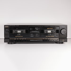 Puede incluir: Una pletina de casete estéreo Aiwa negra con dos compartimentos para cintas etiquetados como Deck 1 y Deck 2. El panel frontal incluye un botón de encendido, una perilla de nivel de grabación, un conector para auriculares y varios botones.