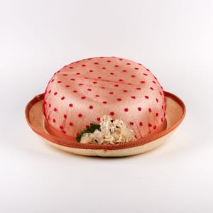 Puede incluir: Un sombrero redondo con una copa de color melocotón claro cubierta por una malla de red con lunares rojos. El sombrero tiene un ala ancha de color claro con un borde color melocotón oscuro y un grupo de pequeñas flores de tela.