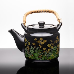 May include: A glossy black ceramic teapot featuring a floral pattern in shades of yellow and green. The teapot has a curved spout, a matching lid with a small knob, and a light-coloured, woven bamboo handle attached to two side loops.