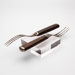 May include: Two vintage forks with dark brown wooden handles and weathered metal tines. Each fork features a metallic bolster and a rounded end cap. The metal surfaces show signs of age and oxidation, contrasting with the dark, textured wood.