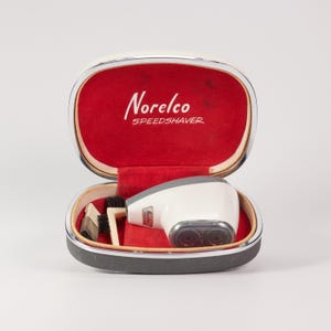 May include: A vintage Norelco Speedshaver electric razor with a white body and dual rotary heads, housed in an oval carrying case with a red velvet interior. The case lid features the white text Norelco SPEEDSHAVER. Includes a small cleaning brush.
