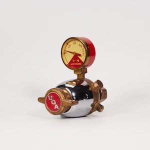 May include: A chrome and brass acetylene gas regulator featuring a round gauge with a red and cream face. The gauge displays markings for 1/4, 1/2, 3/4, and FULL, with the word ACETYLENE printed below the needle. A red circular knob marked UGA is present.