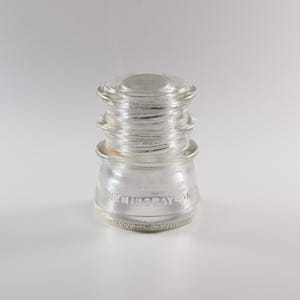 May include: A clear glass electrical insulator with a tiered, cylindrical shape. The base features the embossed text HEMINGRAY-16. The object has a smooth, rounded top and a wider, flared bottom edge with a textured, slightly rough finish.