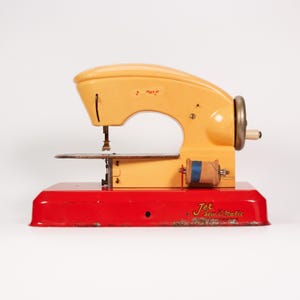 May include: A vintage Jet Sew-O-Matic sewing machine featuring a yellow upper body and a bright red base. The machine includes a metal hand wheel, a needle assembly, and a wooden spool with blue thread attached to the side of the yellow frame.