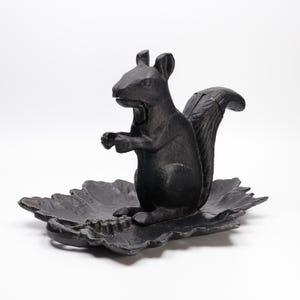 May include: A black cast iron nutcracker shaped like a squirrel standing on its hind legs. The squirrel is positioned on a base molded in the shape of a textured leaf. The metal surface has a matte finish and shows detailed contours of the animal.