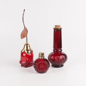 May include: Three red glass decorative items: a rose-shaped bottle with a gold-tone metal stem and leaf, a small round bottle with a gold-tone cap and textured pattern, and a tall bottle with a diamond-cut base and a cork stopper.