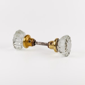 May include: A door knob set featuring two clear, fluted glass knobs attached to brass-toned metal bases. The two knobs are connected by a central metal spindle, with each base showing a small screw hole for installation on a door surface.