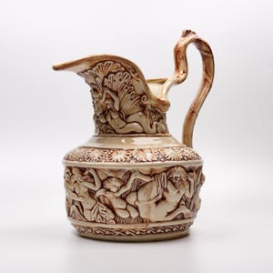 May include: A cream-colored ceramic pitcher featuring intricate brown-toned relief carvings of mythological figures. The vessel has a wide spout, a curved handle, and a detailed band of floral motifs encircling the upper and lower sections.