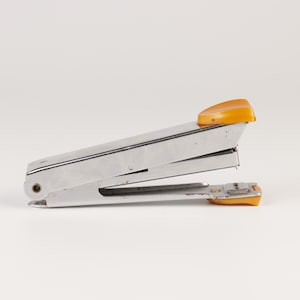 May include: A metal stapler featuring a silver-toned body with a curved, bright yellow plastic cap on the top lever and a matching yellow plastic base component at the front, designed for fastening paper documents with a standard mechanical hinge.