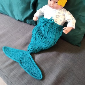 Può includere: Una bambola di un bambino che indossa un costume da coda di sirena in crochet turchese con un cerchietto in crochet giallo. La bambola è sdraiata su un divano grigio con cuscini turchesi.