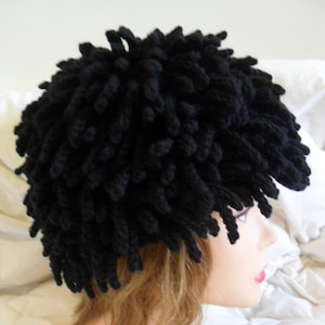 Può includere: Un cappello all'uncinetto nero con un design soffice e testurizzato. Il cappello è indossato sulla testa di una persona.