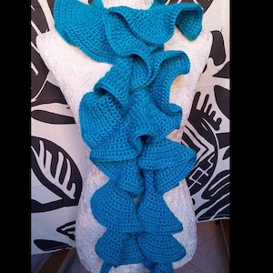 May include: A turquoise crochet scarf with a ruffled design is displayed on a mannequin. The scarf is made of a soft, textured yarn and features a series of cascading ruffles. The words "Crochet Tutorial" are at the top of the image.