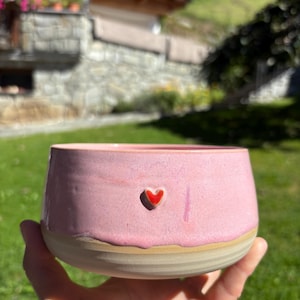 Könnte beinhalten: Eine Keramikschale mit einem glatten, hellrosa glasierten oberen Teil und einem natürlichen, unglasierten beigen Boden. Ein kleines, erhabenes rotes Herz ist mittig auf der rosa Außenseite angebracht. Die Schale hat eine weite Öffnung.