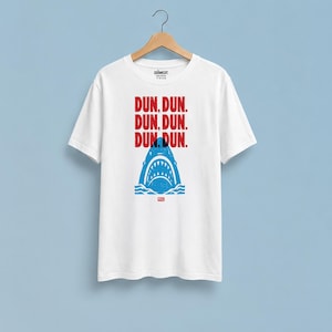 Op de afbeelding: Een wit T-shirt met korte mouwen en een blauwe afbeelding van een haai die uit golven komt. Boven de haai staat de tekst "DUN, DUN. DUN, DUN. DUN, DUN." in het rood. Het T-shirt hangt aan een houten kledinghanger tegen een blauwe achtergrond.