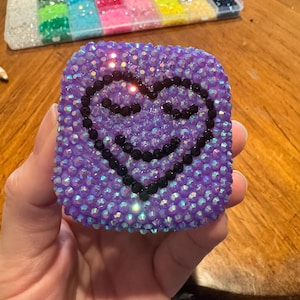 Puede incluir: Un objeto de forma cuadrada cubierto de pedrería púrpura iridiscente con un diseño de corazón central delineado en pedrería negra. El corazón incluye dos ojos cerrados y una sonrisa curva, creados con pequeñas piedras oscuras y brillantes.